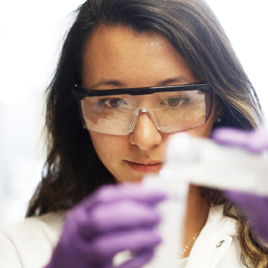Photo of a female researcher wearing goggles and putting liquid into a test tube.