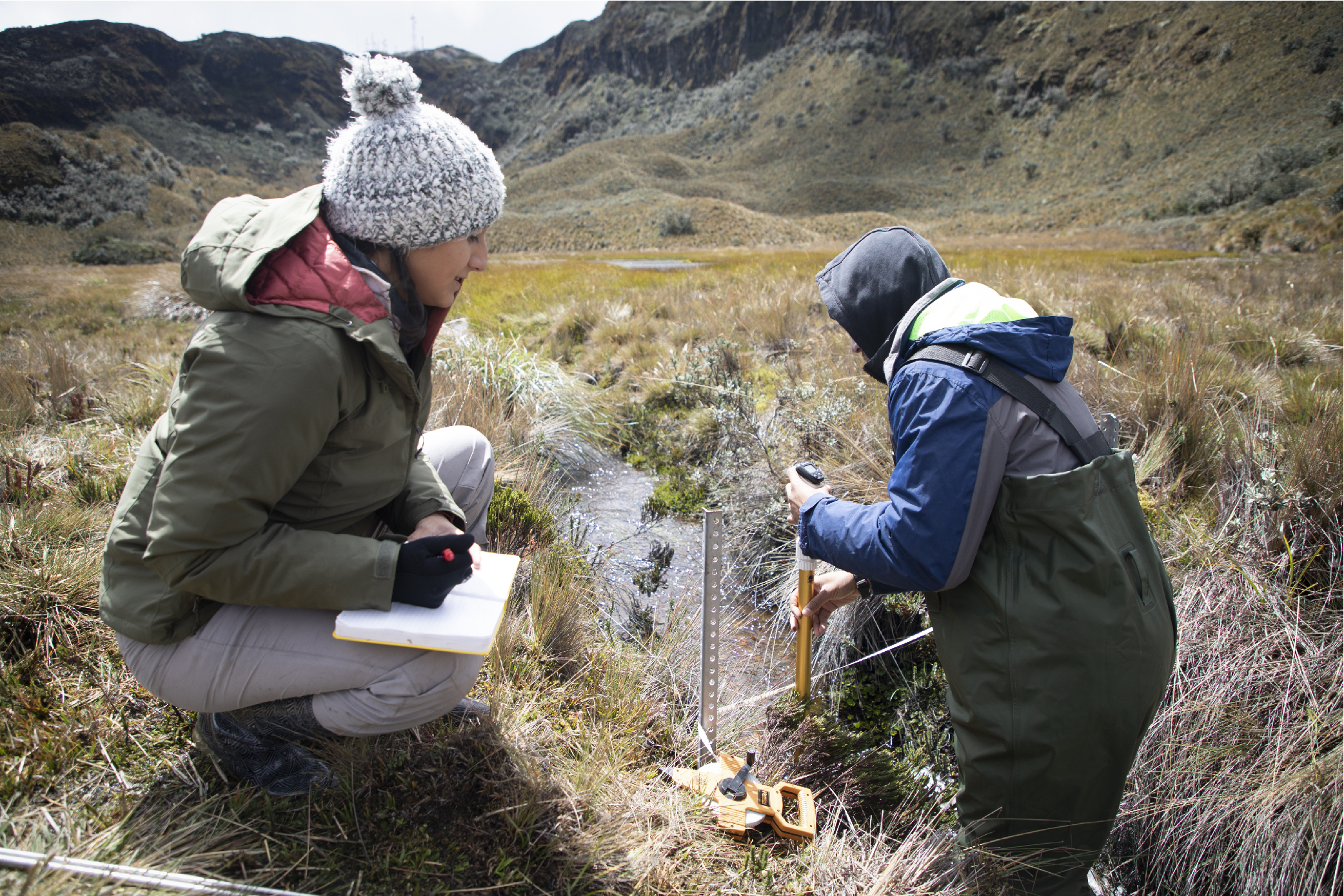 Maribel Herrera sits along a stream in Ecuador and records data from a water sensor being monitored by Nehemiah Stewart.