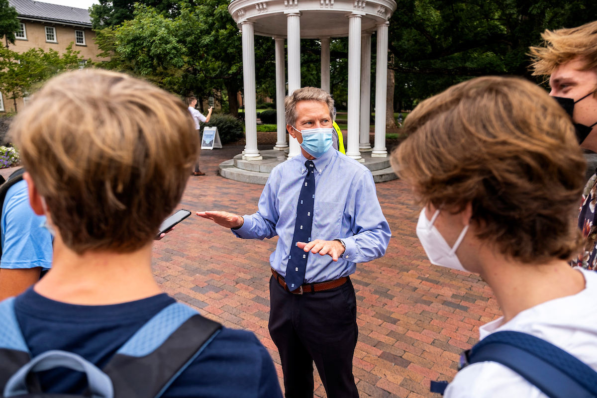 Photo of Kevin Guskiewicz, masked, talking with students in front of the old well.