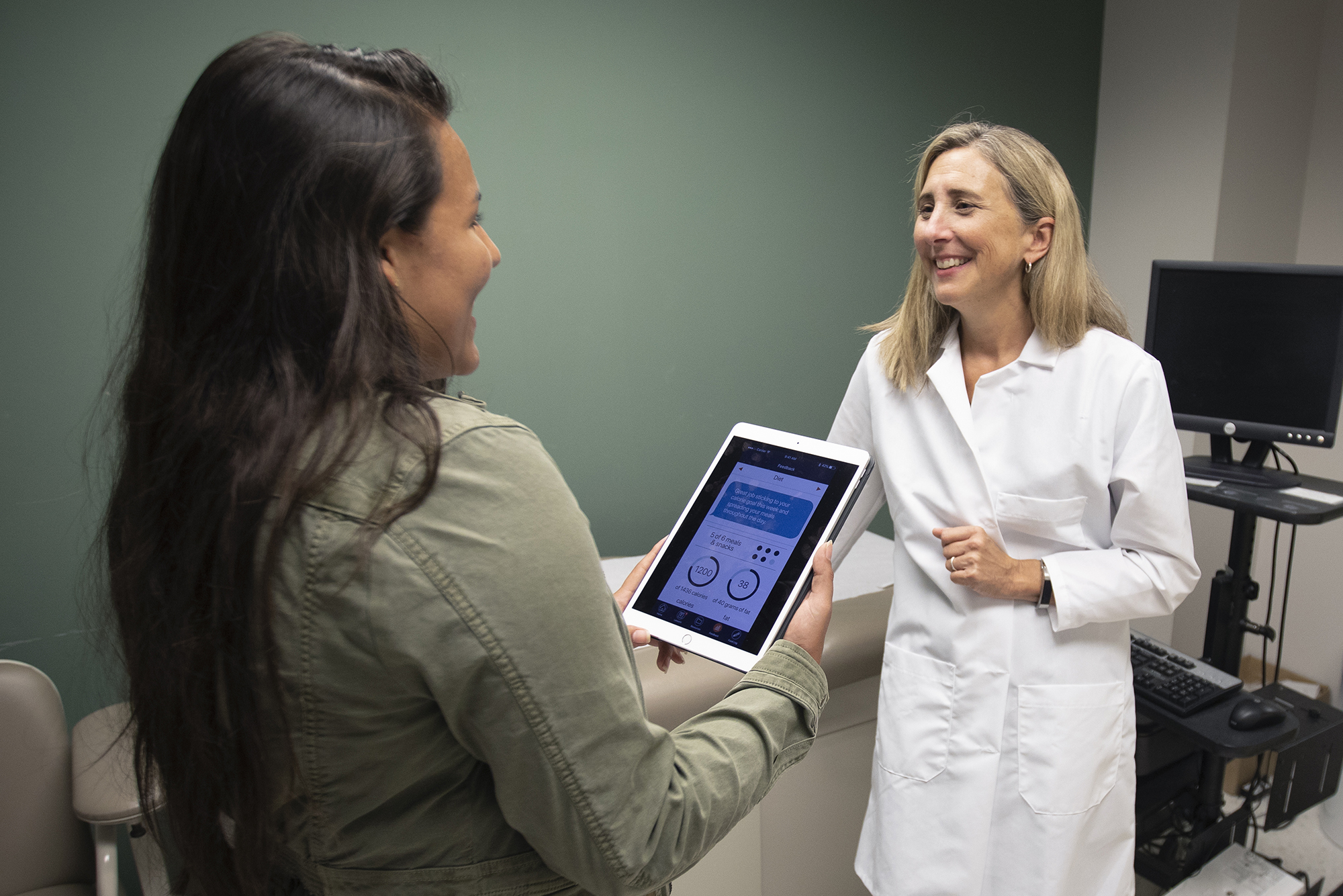 Doctor talks to a patient while they hold an iPad, displaying an app.