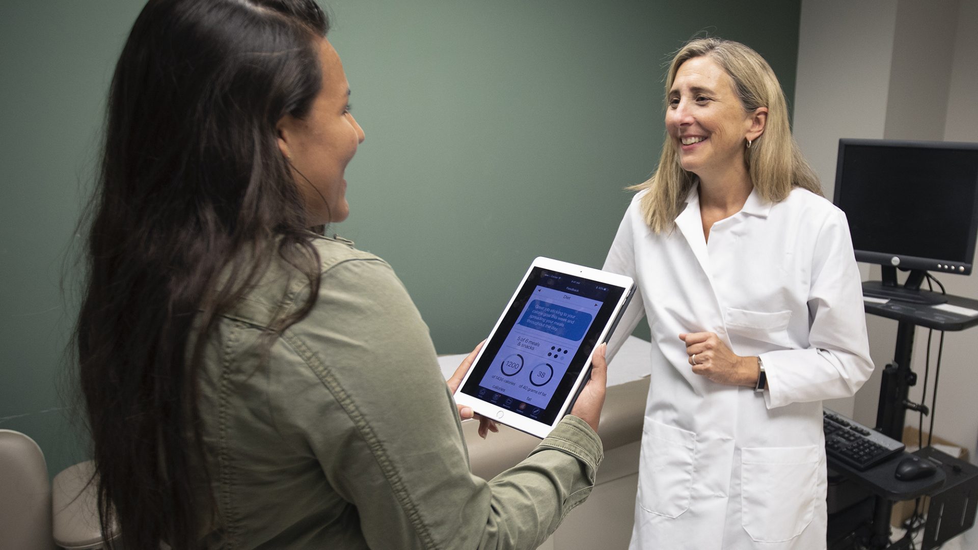 Deb Tate (right) speaks with a young woman holding an iPad with the mPWR app on it in an exam room.