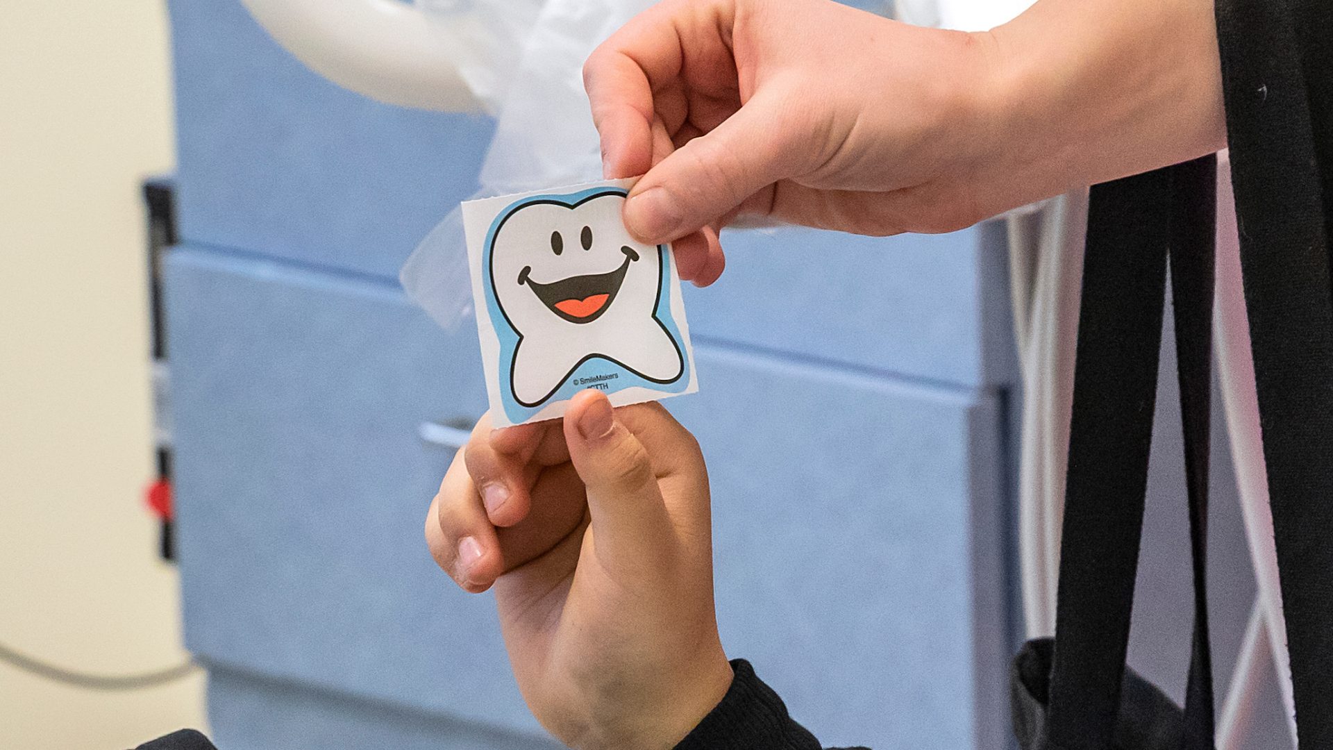 Two people, together, hold a sticker of a tooth cartoon smiling.
