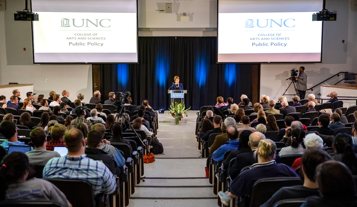 A woman presents a speech behind a podium between two screens that read "UNC College of Arts and Sciences Public Policy" to a room full of people.