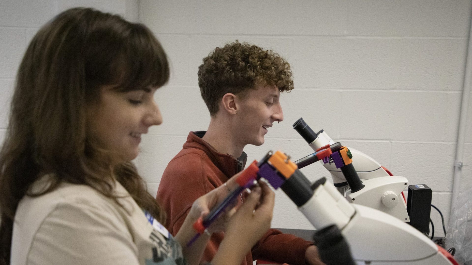 Two students smile as they look into table top microscopes.