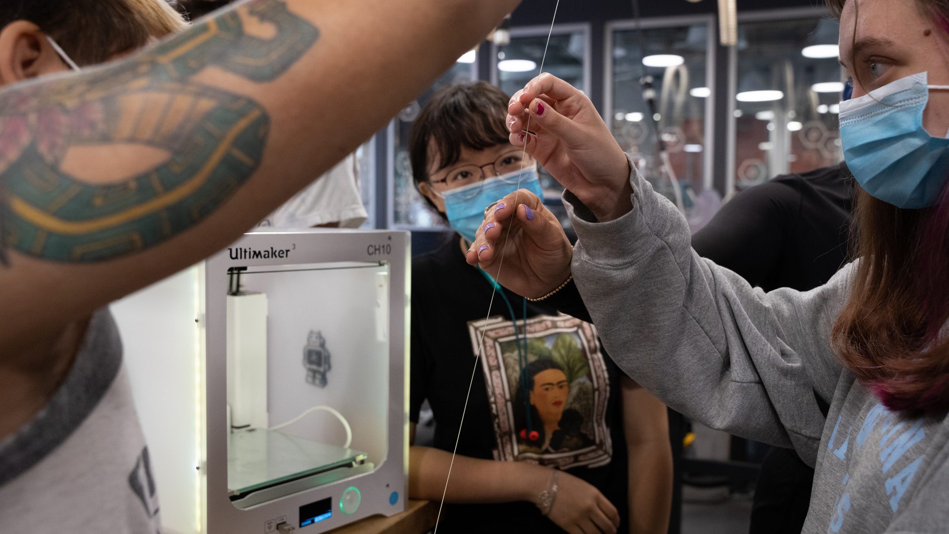Photo of students, in masks, as they hold a thread of plastic filling for the 3D printer in the BeAM Makerspace.