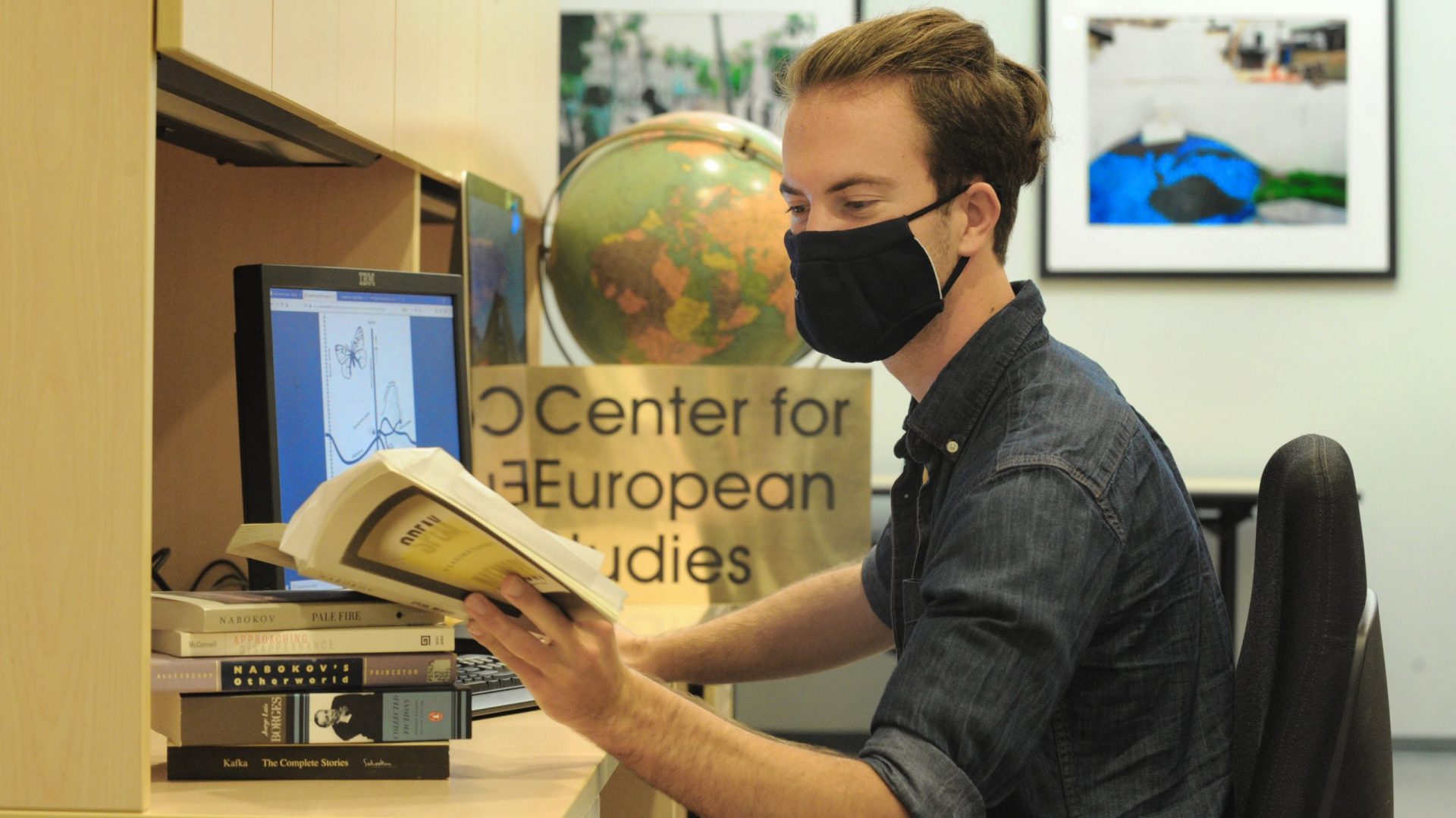 Student, in a mask, reads a book while at a computer.
