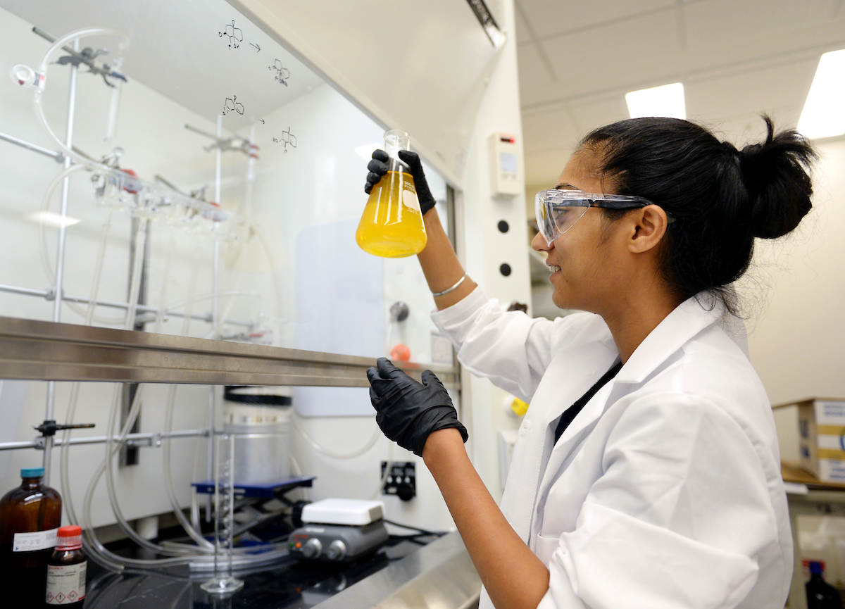 Graduate student holds a chemistry flask full of liquid in their hands, at eye level. They're in a lab setting, wearing a white coat, rubber gloves, and protective glasses.