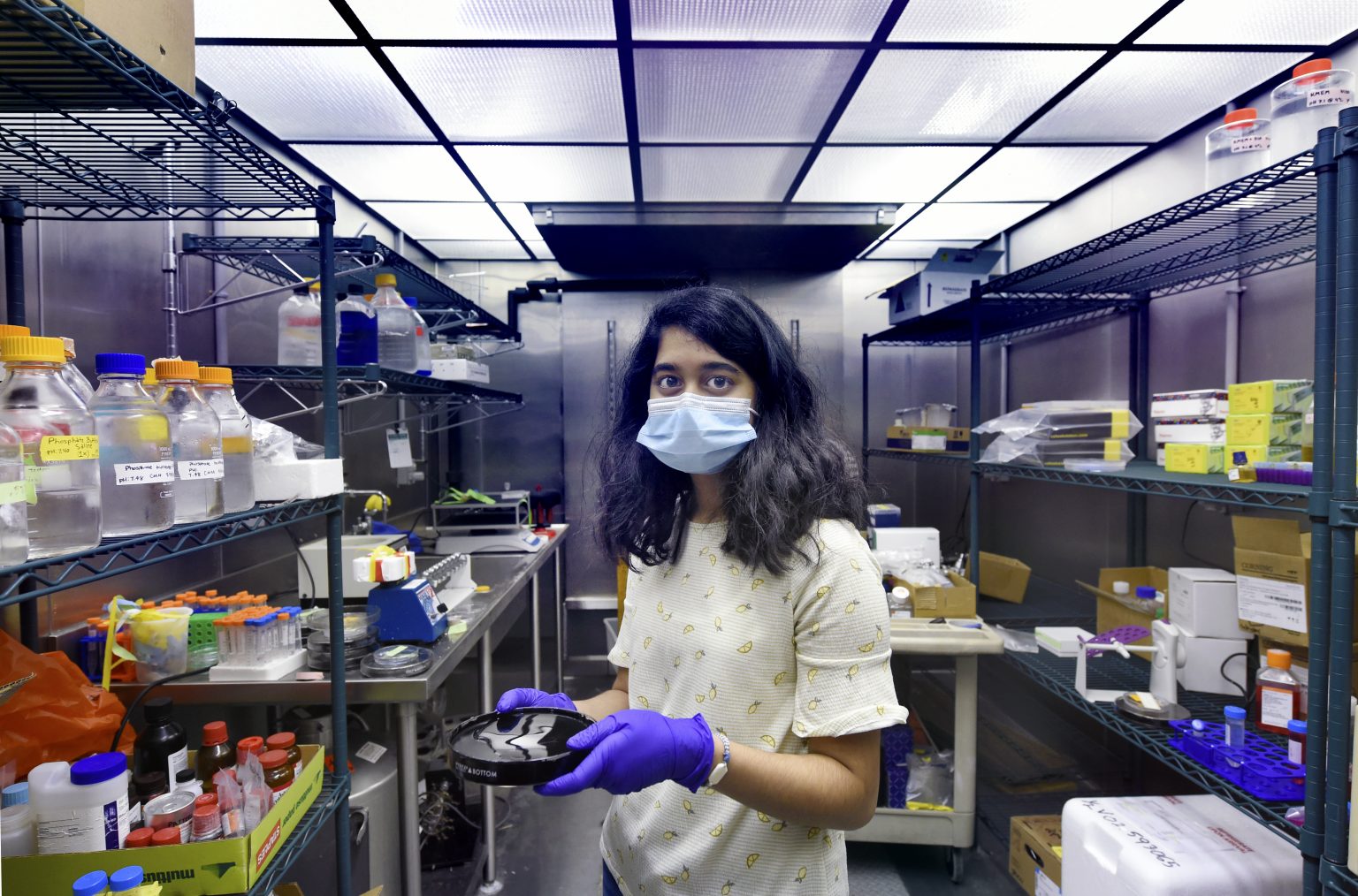 A girl stands in a lab, holding a dish and wearing a face mask.