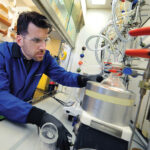 Photo of Frank Leibfarth in the lab wearing protective gear, bent over lab equipment.