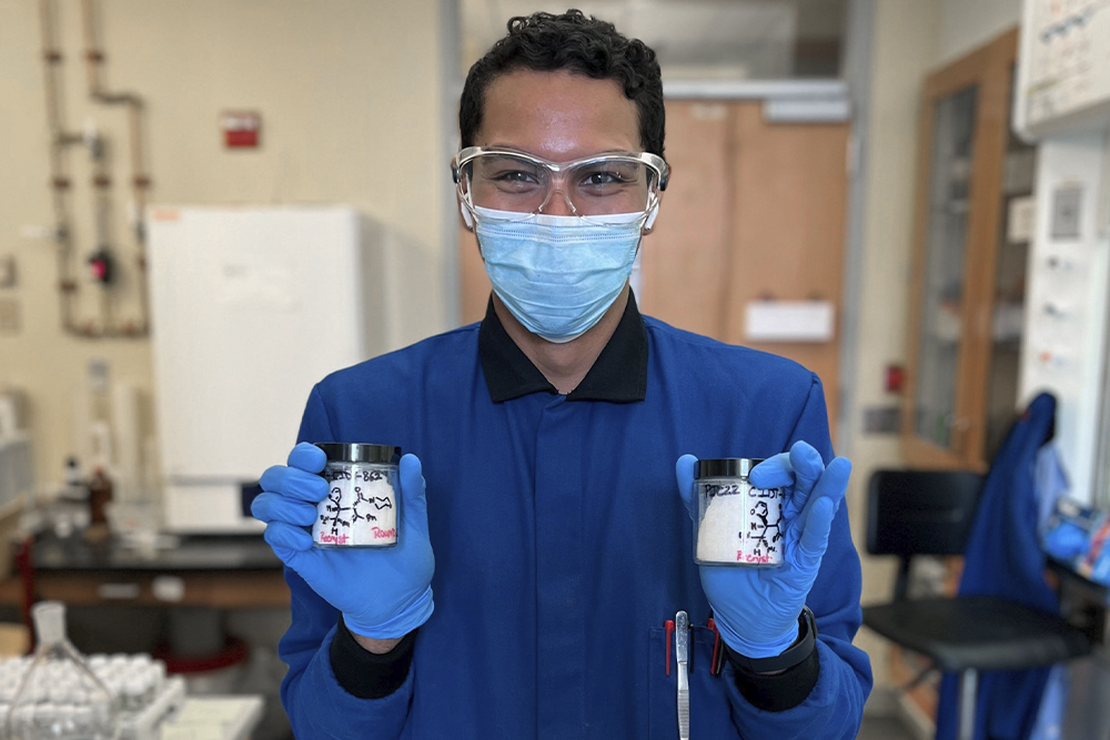 An image of Pedro de Jesús Cruz holding containers of solid compound, dressed in protective lab gear.