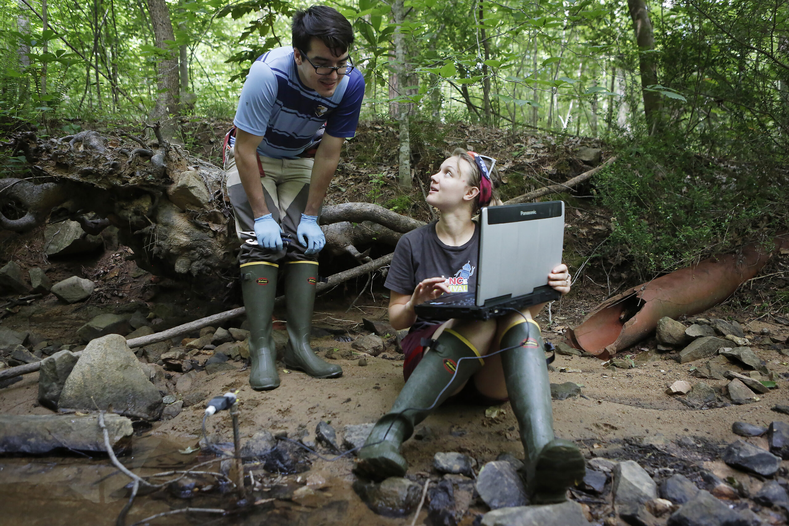 Two researchers sit next to a stream, one with a laptop in their hand, discussing their research.