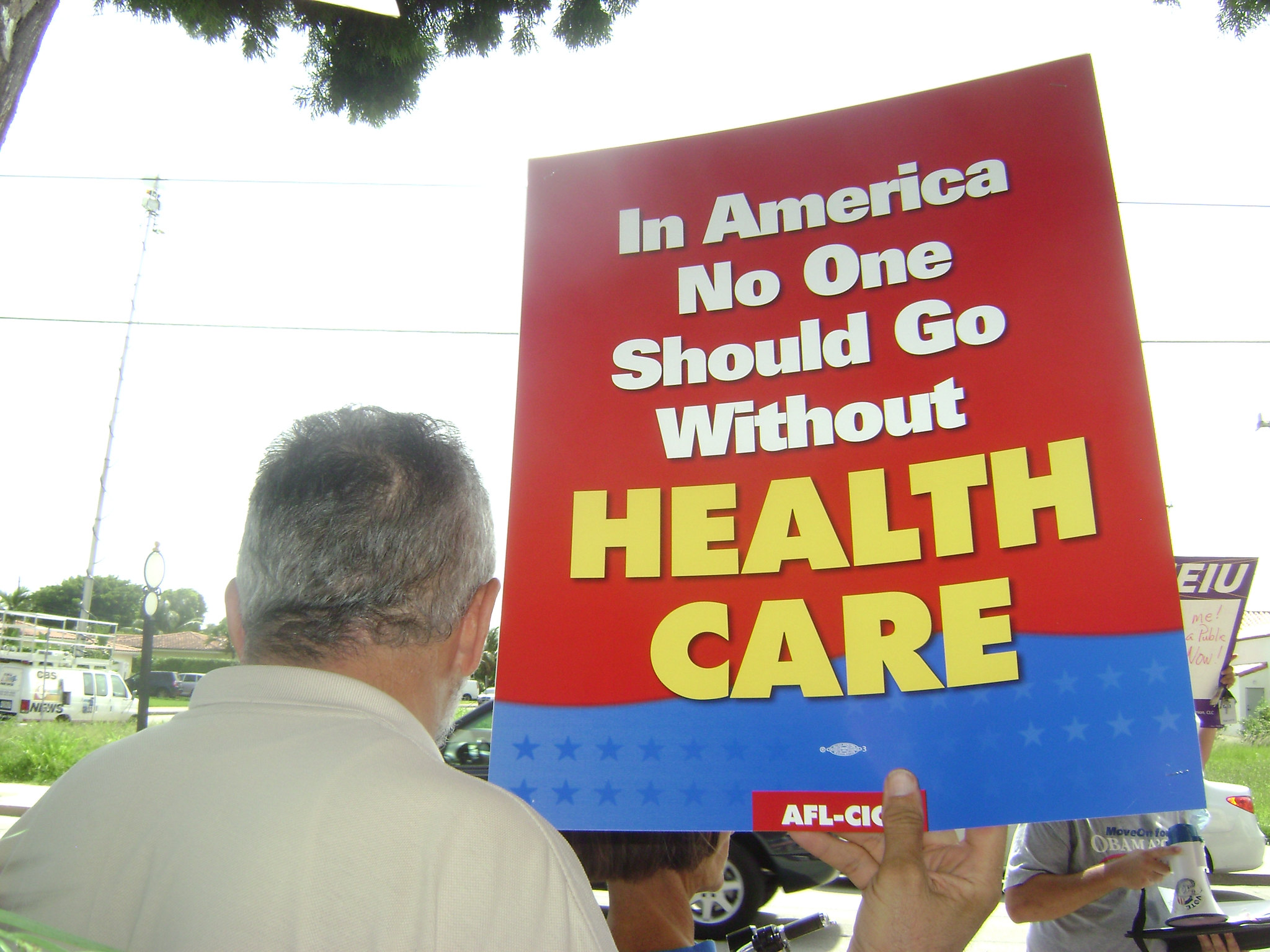 A man holds a sign that says "In America No One Should Go Without Health Care."
