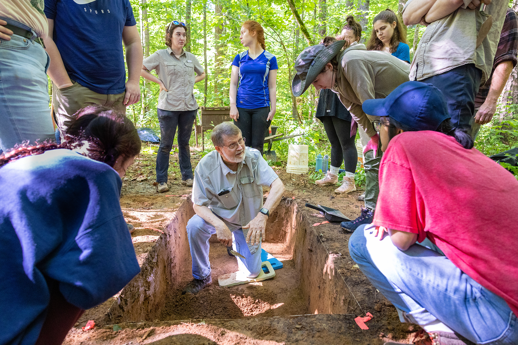 An archaeology professor stands in a hand dug pit, talking to students as the crowd around him.