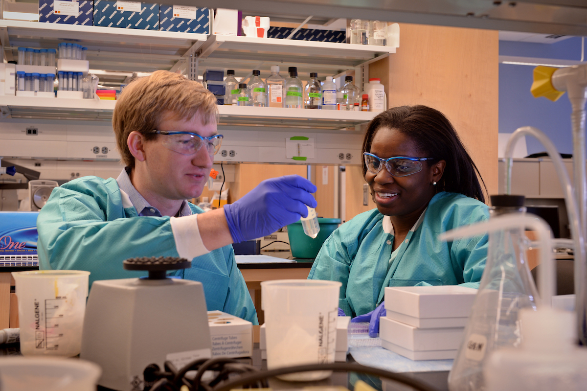 Two researchers, in PPE gear, look at a vial.