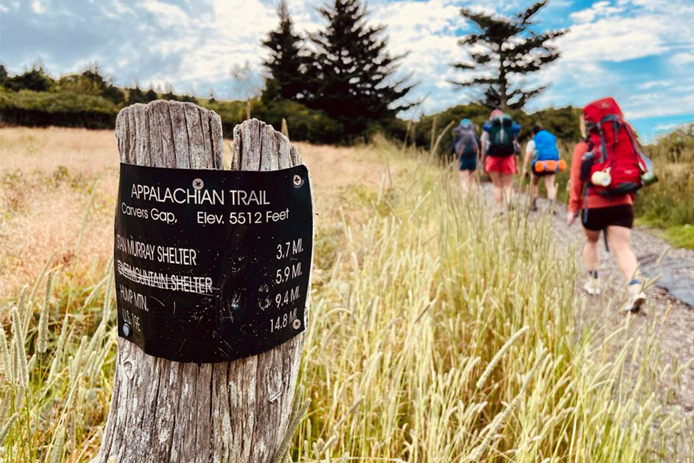 Photo showing an outdoor wooden post that reads "Appalachian Trail" with people backpacking in the background.