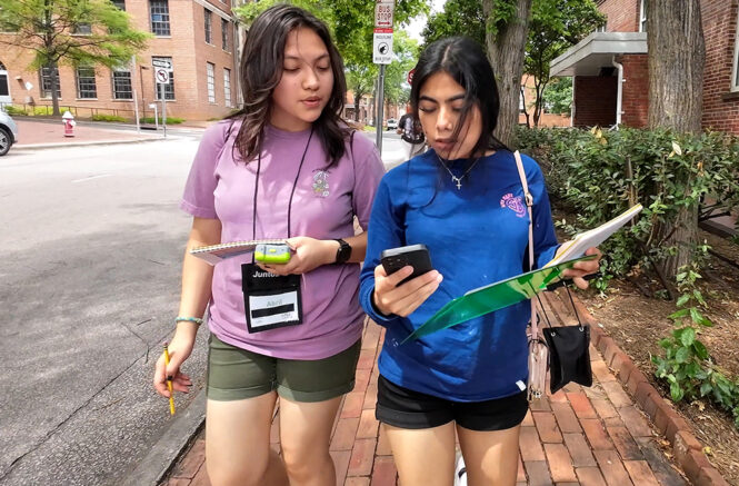 Two high school students look at a monitor as they walk down the street.