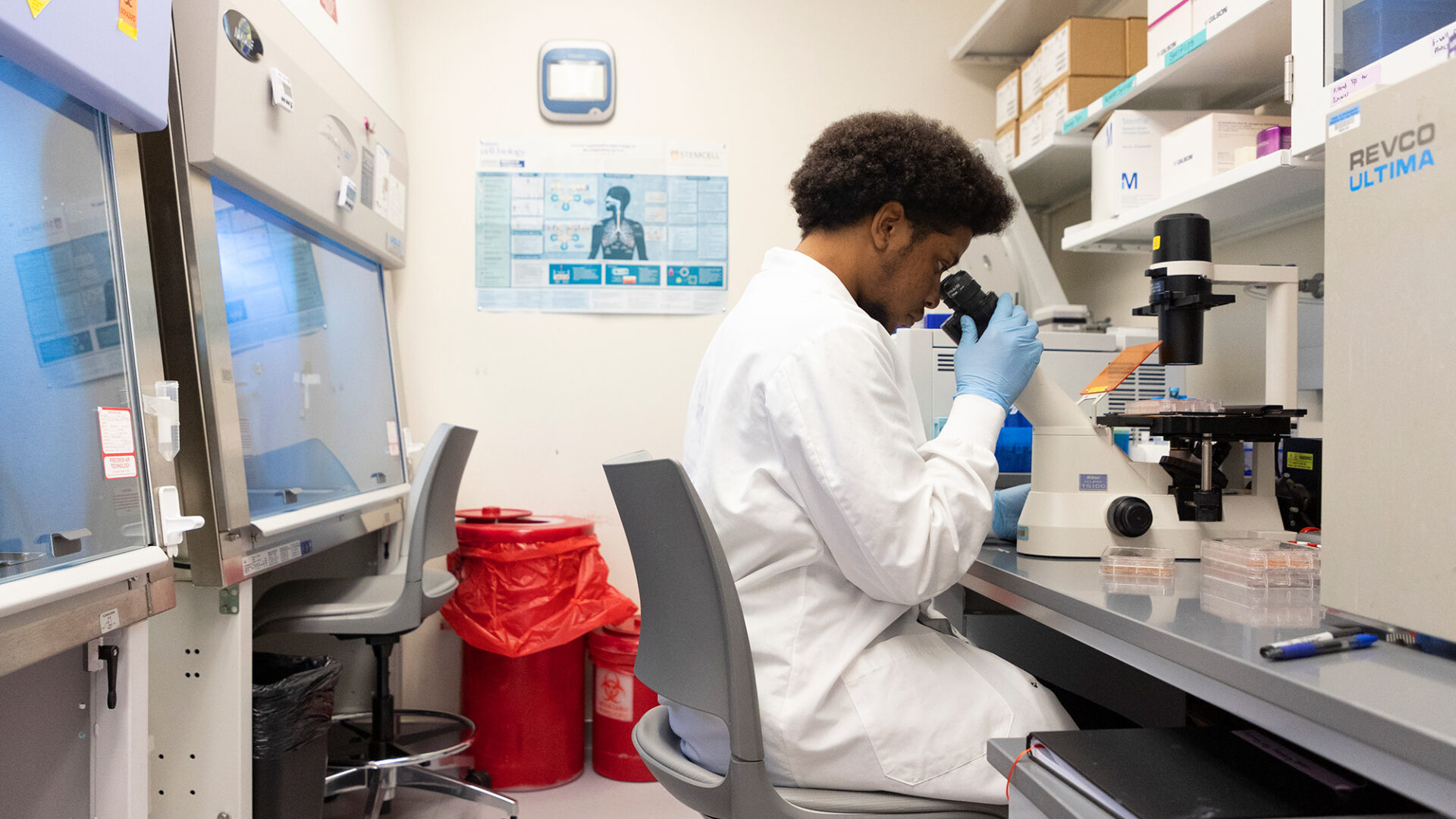 Researcher sites in their lab in a white coat behind a microscope.